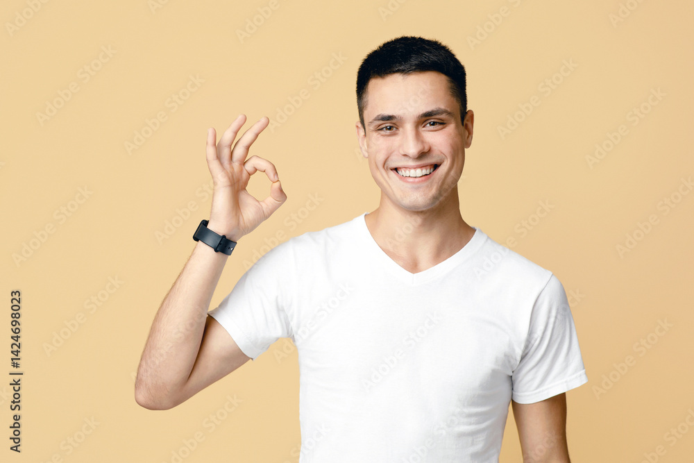 Successful man with gestures celebrating victory. Happy attractive young blogger student in white t-shirt with smart watch shows sign okay, isolated on light sand background, empty space, studio shot Successful man with gestures celebrating victory. Happy attractive young blogger student in white t-shirt with smart watch shows sign okay, isolated on light sand background, empty space, studio shot