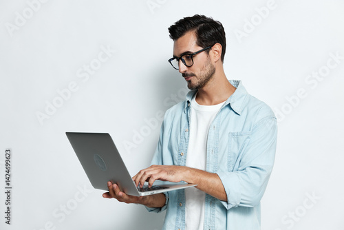 young man with laptop wearing glasses and denim shirt standing isolated on light background working on computer in casual style