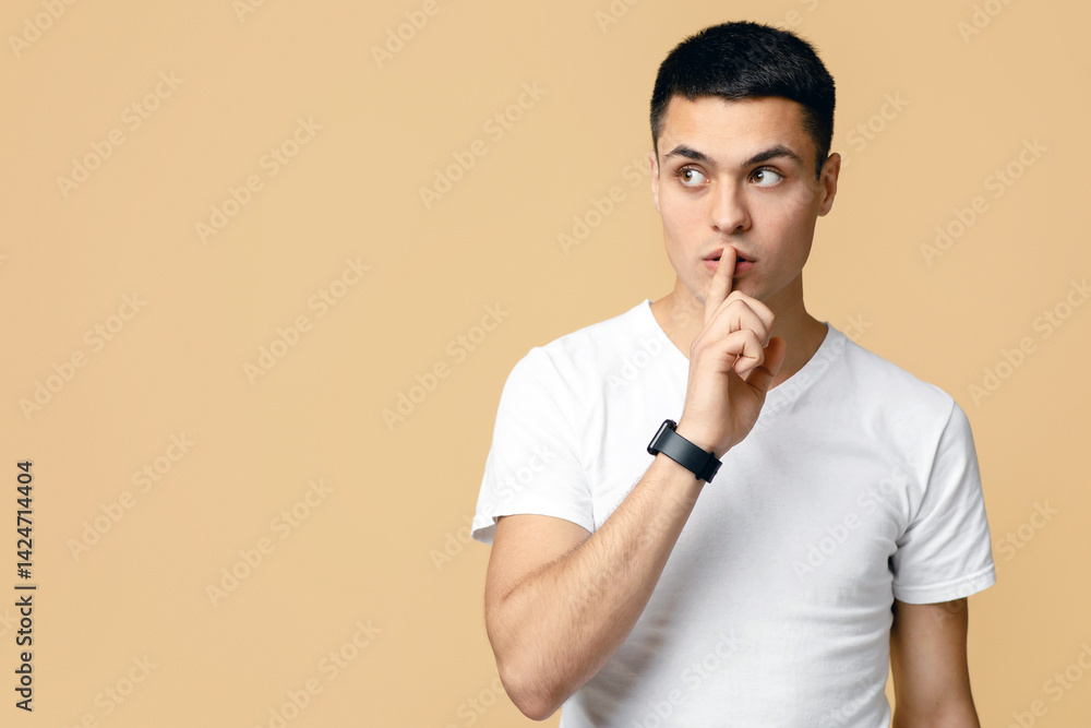 Keep secret, be silent, great offer and advertising. Serious young blogger student man in white t-shirt with smart watch makes secret sign, isolated on light sand background, empty space, studio shot Keep secret, be silent, great offer and advertising. Serious young blogger student man in white t-shirt with smart watch makes secret sign, isolated on light sand background, empty space, studio shot