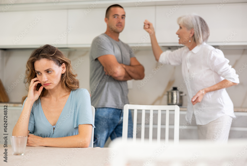Fototapeta premium Young girl is sitting at table in kitchen quarreling with her mother-in-law and husband