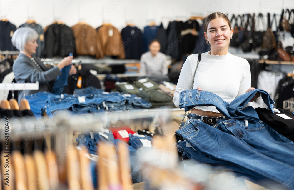 Glad young female customer looking at jean trousers put on stand in clothes store