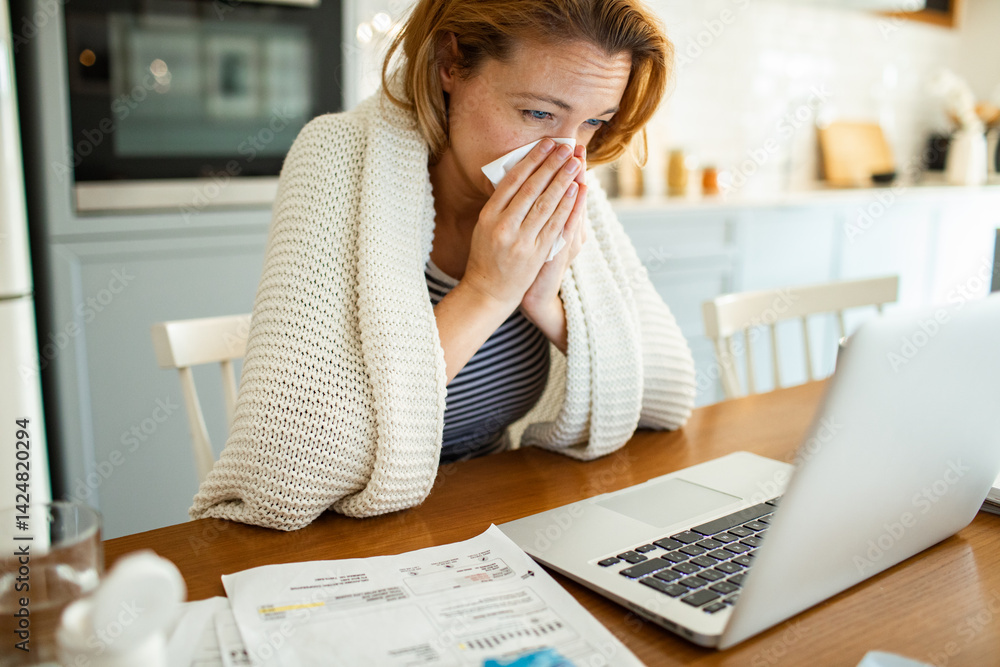 Sick woman blowing nose in tissue at kitchen table with laptop