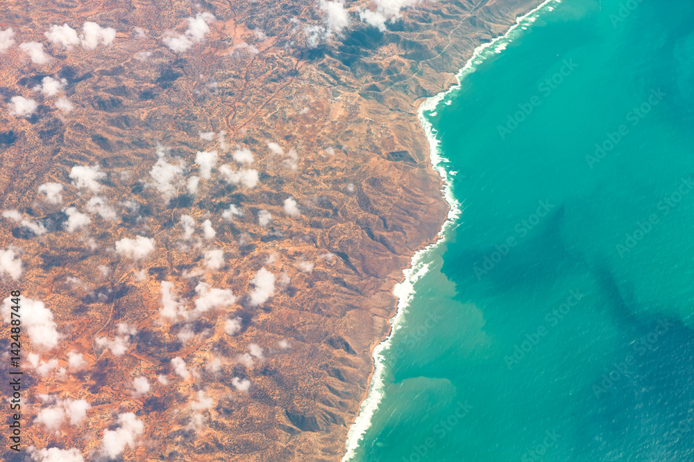 Aerial view captures a rugged, mountainous coastline meeting deep blue ocean under a partly cloudy sky. Contrast between arid, rocky terrain and vibrant sea highlights dramatic beauty of landscape