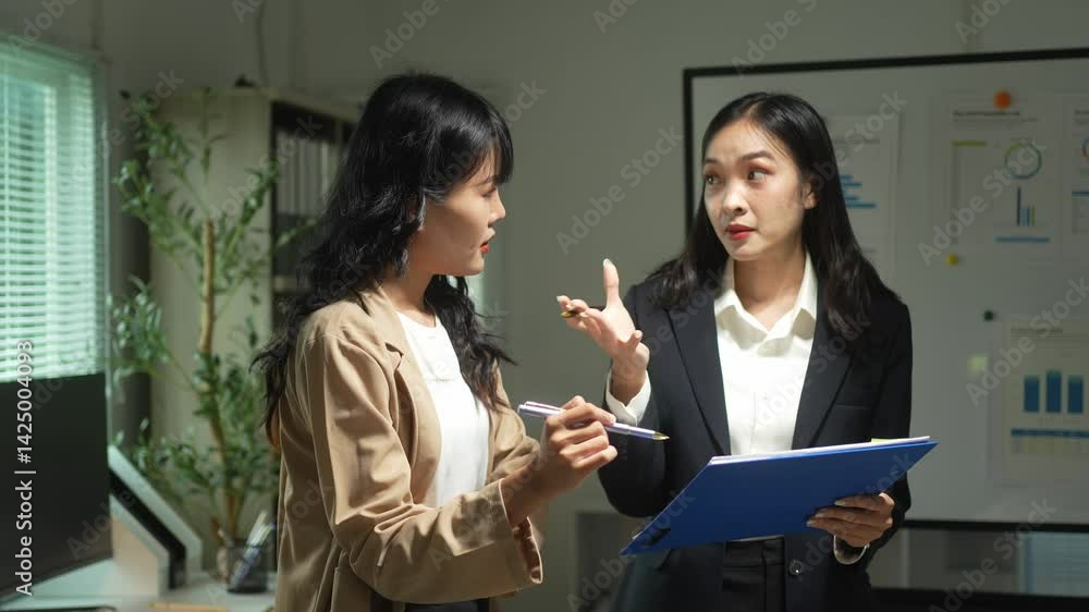 Two women are talking in a room with a white board. One of them is holding a blue clipboard Two women are talking in a room with a white board. One of them is holding a blue clipboard