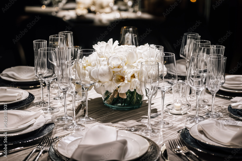 The image shows a elegantly set dining table with white flowers as the centerpiece, surrounded by crystal glasses, white plates, and neatly folded napkins