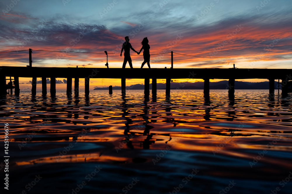The image shows a silhouette of a couple holding hands and walking on a pier at sunset