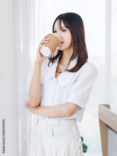Smiling asian young beautiful pretty happy woman standing near curtains window in bedroom at home holding coffee cup in morning and enjoying sunlight. Attractive female wearing white shirt white pants