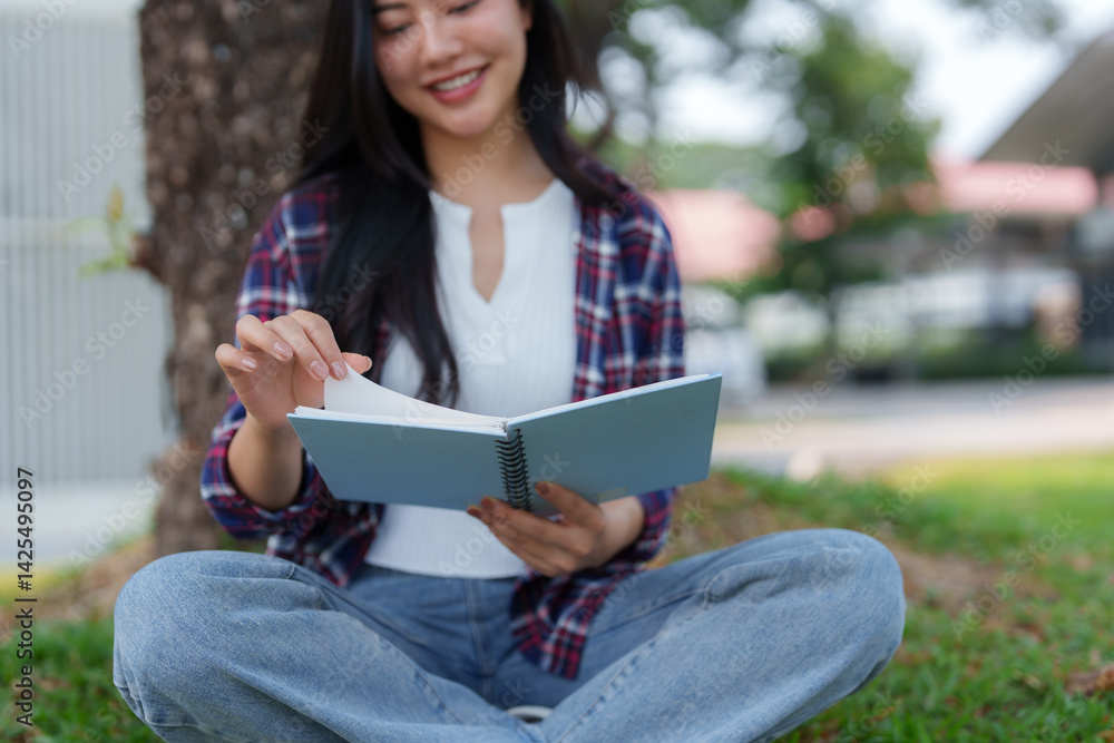Fototapeta premium Young asian woman enjoys reading a book while sitting on grass in a park, turning a page with a smile, showcasing the joy of learning and leisure