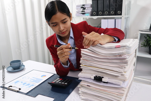 A young professional woman, examining a tall stack of documents with a focused expression.background are filled with more documents and black binders, suggesting a busy office environment