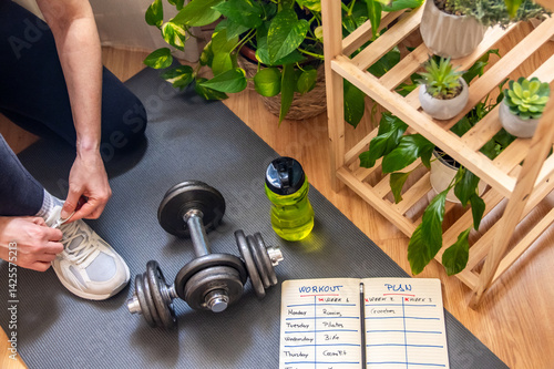 Woman tying her running shoes, preparing for a home workout session on a fitness mat with dumbbells and a workout plan, surrounded by houseplants promoting wellness and a healthy lifestyle