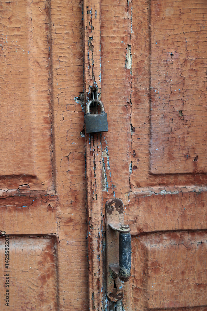 Close Up Vertical View Of An Old Wooden Door With Heavily Cracked And Peeling Brown Paint, Metal Handle And A Padlock. Texture Of Decay And Neglect. Locked Concept. Close Up Vertical View Of An Old Wooden Door With Heavily Cracked And Peeling Brown Paint, Metal Handle And A Padlock. Texture Of Decay And Neglect. Locked Concept.