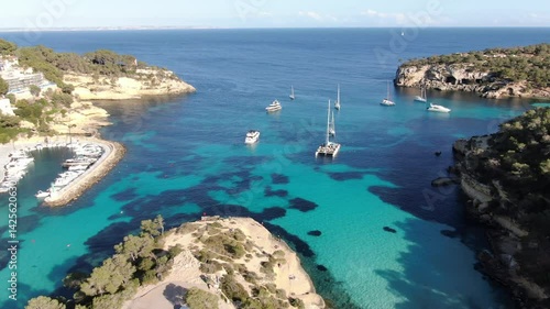  Aerial view of luxury yachts located in a small bay on the coast of Mallorca.  The bay is  filled with several boats and yachts, some anchored and others moving slowly across the turquoise-blue water