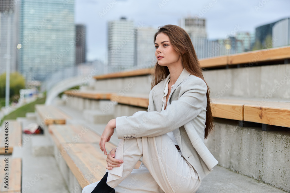 Young woman in light suit sitting thoughtfully on urban steps with modern cityscape in background, expressing contemplation, fashion, and business style