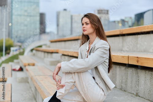 Obraz na płótnie Young woman in light suit sitting thoughtfully on urban steps with modern cityscape in background, expressing contemplation, fashion, and business style w salonie