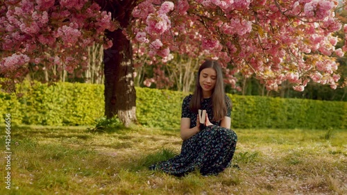 Young woman looks up at the sky with a peaceful expression, holding her Bible. Surrounded by blooming sakura trees, she reflects on faith and gratitude, embracing a moment of spiritual connection.