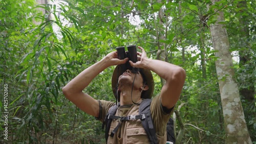 An Asian male traveler explores the forest in search of birds, gazing into the treetops through binoculars, embracing eco-tourism and a mindful connection with nature.