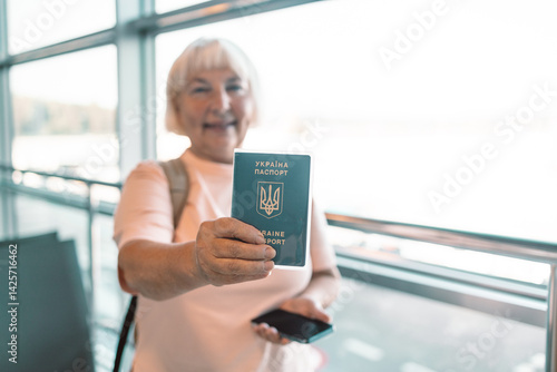 Obraz na płótnie Senior woman stands in an airport terminal, smiling and holding her passport and smartphone. She is ready for her flight, with modern airport windows and a bright, welcoming atmosphere in the w salonie