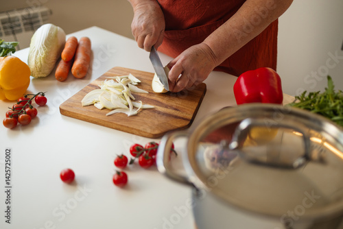 A person slices onion on a wooden cutting board in a bright kitchen. Various fresh vegetables, including tomatoes, carrots, and bell peppers, are placed around for meal preparation