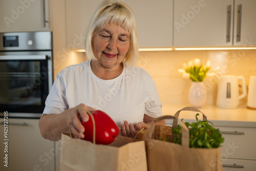 An elderly woman is unpacking groceries from paper bags in her kitchen. She places a jar of food and fresh produce on the counter, enjoying the convenience of home delivery