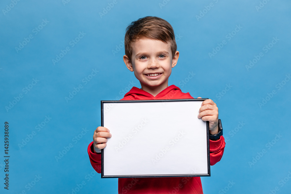 Portrait of happy laughing little boy holding white drawing board on blue background, young boy is holding a drawing board in his hands. Mock up. Copy space. High quality photo