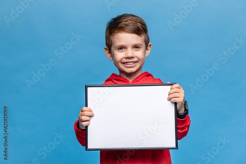 Obraz na płótnie Portrait of happy laughing little boy holding white drawing board on blue background, young boy is holding a drawing board in his hands. Mock up. Copy space. High quality photo w salonie