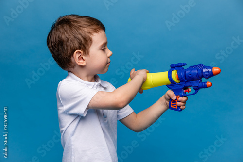 Obraz na płótnie Laughing child boy playing with water gun toy on blue background. Pool toys and water fun for family with children. Summer vacation with kids. Active outdoor sport for preschooler w salonie