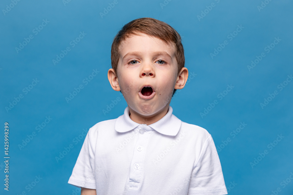 Portrait of scared little boy wearing white t-shirt making a grimace with his mouth open. Child is very surprised on blue background. Mock up. Shock, amazement, surprise concept.