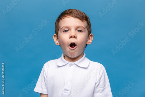 Obraz na płótnie Portrait of scared little boy wearing white t-shirt making a grimace with his mouth open. Child is very surprised on blue background. Mock up. Shock, amazement, surprise concept. w salonie