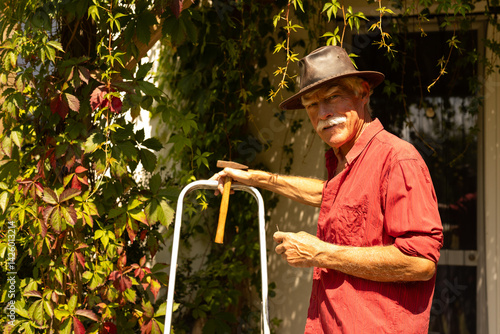 A handsome elderly gentleman in retirement doing small renovation work in his summer cottage. He is standing on the porch with a hammer in his hand next to a ladder. Stylish hat on his head.