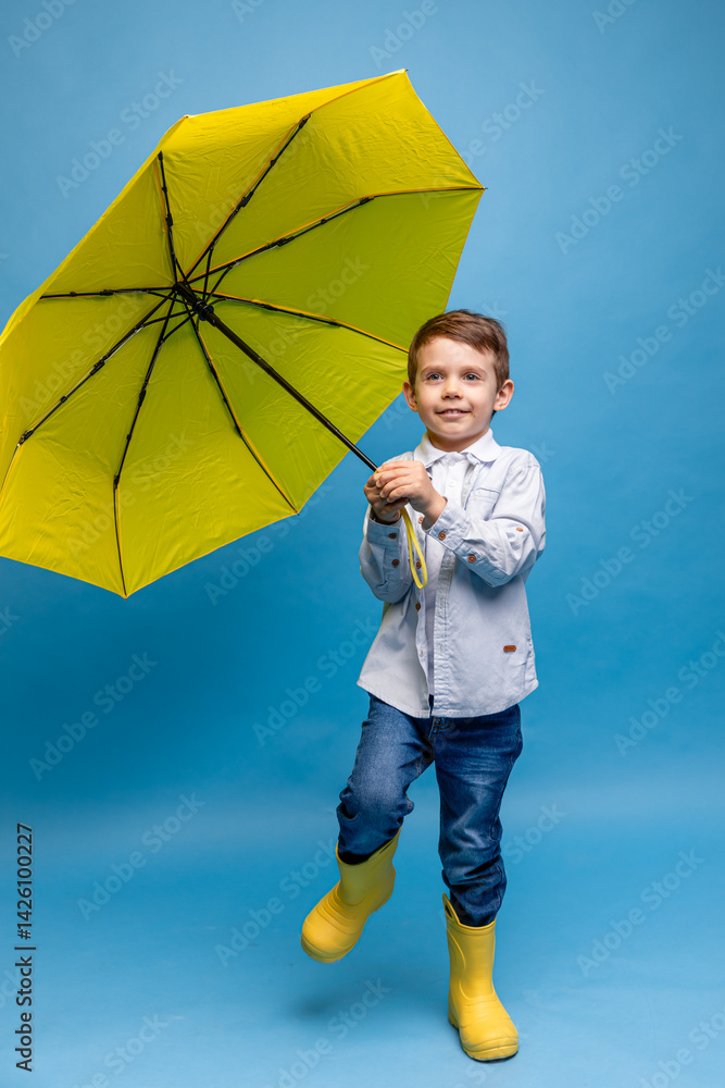 Happy funny smiling child, little boy, with yellow umbrella posing on blue background. Kid is wearing yellow rubber boots. Rainy spring or autumn weather. Clothes for the rain. Copy space