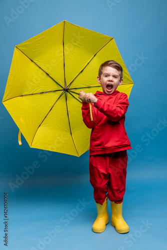 Obraz na płótnie Happy funny smiling child, little boy in red sport suit with yellow umbrella posing on blue background. Kid is wearing yellow rubber boots. Rainy spring or autumn weather. Copy space w salonie