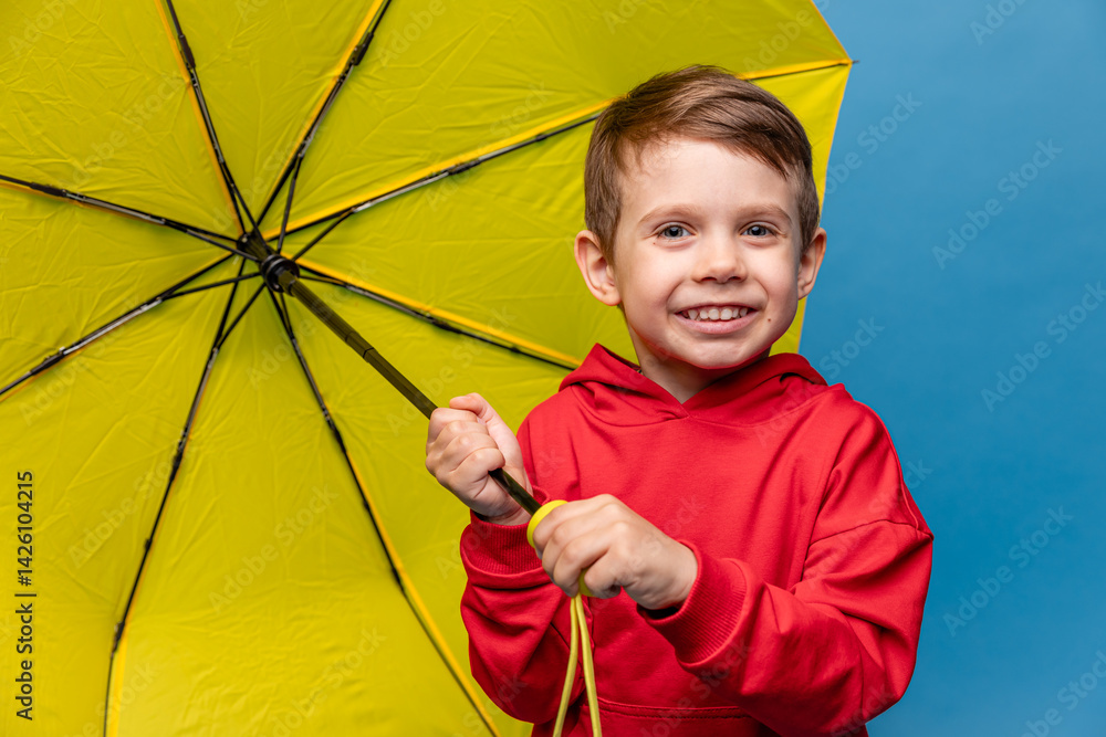 Happy funny smiling child, little boy in red sport suit with yellow umbrella posing on blue background. Kid is wearing yellow rubber boots. Rainy spring or autumn weather. Copy space