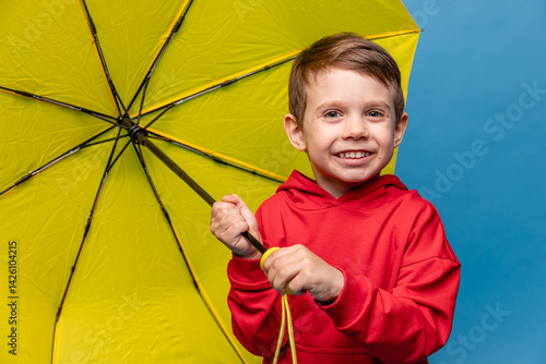 Obraz na płótnie Happy funny smiling child, little boy in red sport suit with yellow umbrella posing on blue background. Kid is wearing yellow rubber boots. Rainy spring or autumn weather. Copy space w salonie
