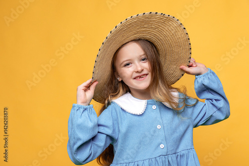 Smiling little child blonde hair girl in blue dress and straw hat on yellow studio background. Summer travel vacation with children, advertising concept. Banner, mock up