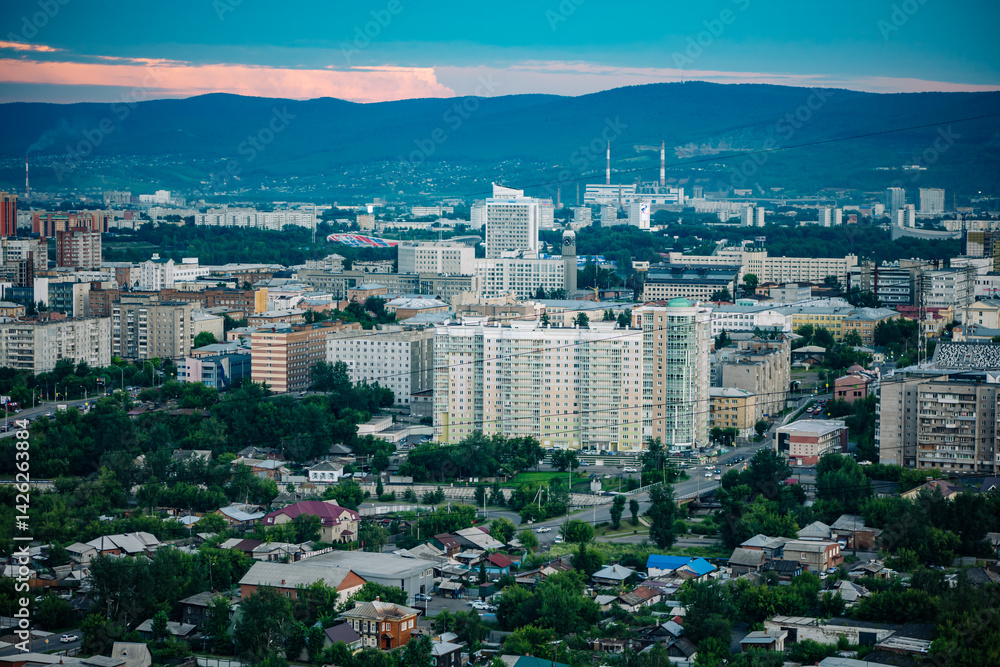 Krasnoyarsk cityscape with residential and industrial buildings at dusk, featuring mountains in the background Krasnoyarsk cityscape with residential and industrial buildings at dusk, featuring mountains in the background