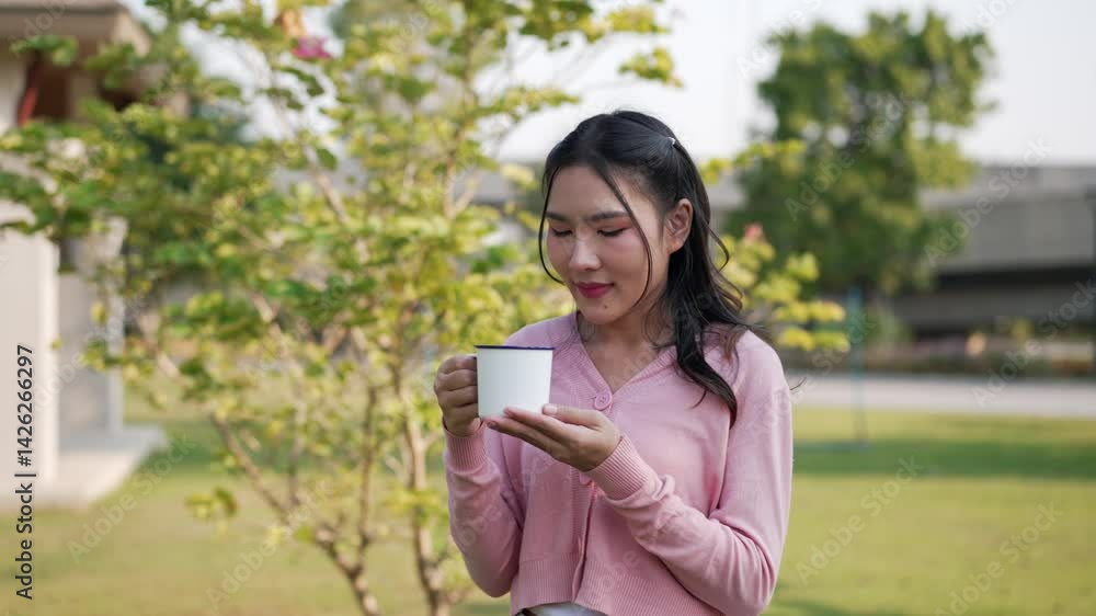 Locked medium shot, Happy Young Asian woman stand and holding warm drink, She smelled the aroma and drank coffee happily during relaxation at outdoor Locked medium shot, Happy Young Asian woman stand and holding warm drink, She smelled the aroma and drank coffee happily during relaxation at outdoor