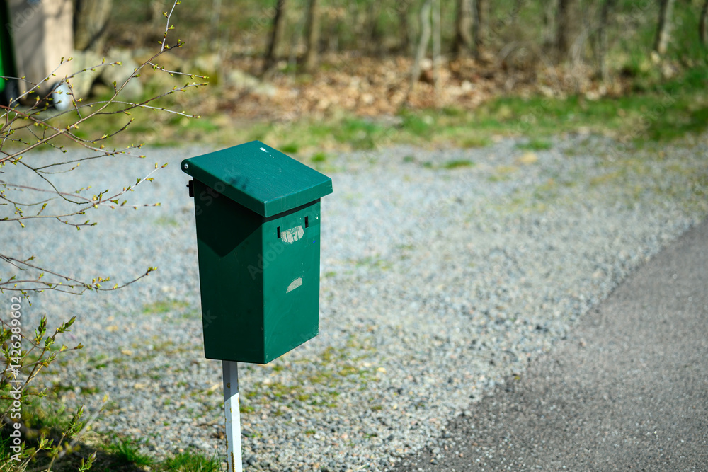 A green mailbox stands prominently on a white post along a gravel driveway. The surrounding area features trees with fresh spring leaves, creating a serene rural atmosphere A green mailbox stands prominently on a white post along a gravel driveway. The surrounding area features trees with fresh spring leaves, creating a serene rural atmosphere