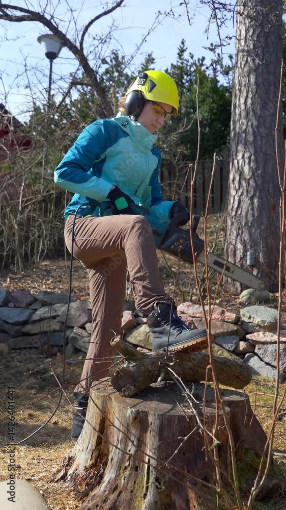 A strong, confident woman sawing through the trunk of an old tree an electric chainsaw, as part of spring cleanup and yard renewal. Dressed in protective gear, she challenges gender norms. A strong, confident woman sawing through the trunk of an old tree an electric chainsaw, as part of spring cleanup and yard renewal. Dressed in protective gear, she challenges gender norms.