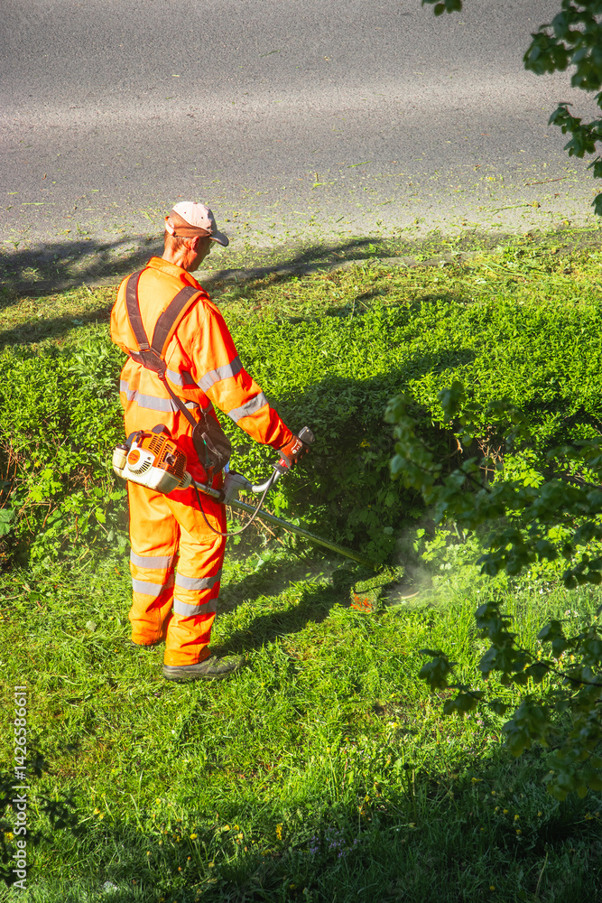 Man in orange uniform and protective glasses mowing green grass with a lawnmower on a sunny day. Bright green lawn and natural outdoor background.