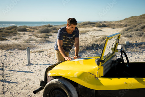 Photos Man leaning and inspecting hood of yellow dune buggy on coastal dune, with ocean