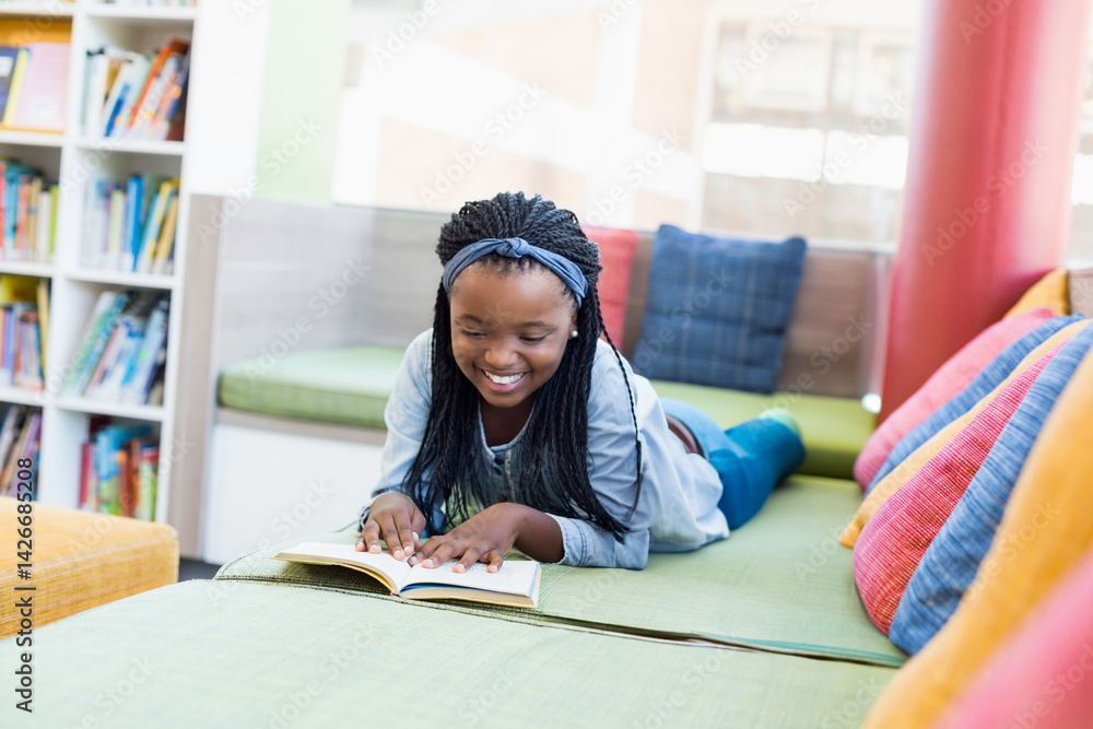 African American teenage girl reading open book on cushioned bench in library nook, smiling
