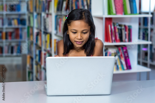 Girl child sitting at white table in library with bookshelves, working on silver laptop