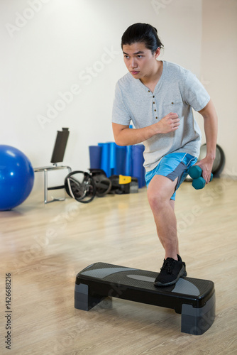 Asian young adult woman stepping onto aerobic step in indoor fitness studio, holding teal dumbbells