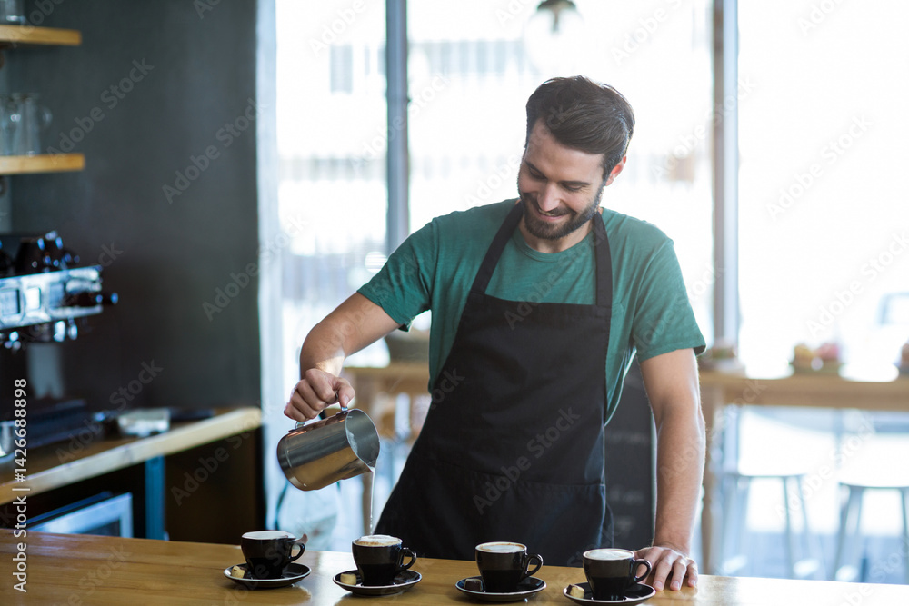 male barista pouring milk with metal pitcher into black cups on counter in coffee shop