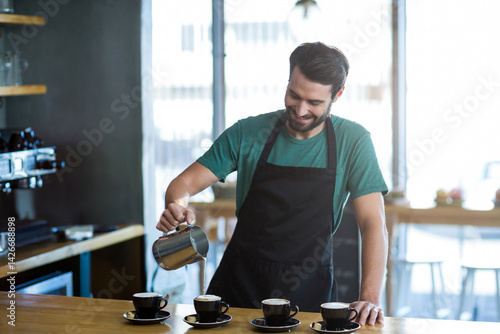 Obraz na płótnie male barista pouring milk with metal pitcher into black cups on counter in coffee shop w salonie