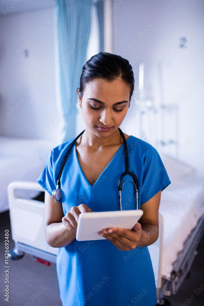 Young Indian nurse standing in hospital room, checking tablet with stethoscope and IV stand