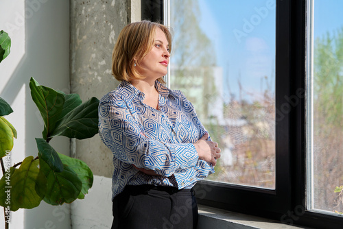 Portrait of beautiful middle-aged woman with crossed arms looking out window