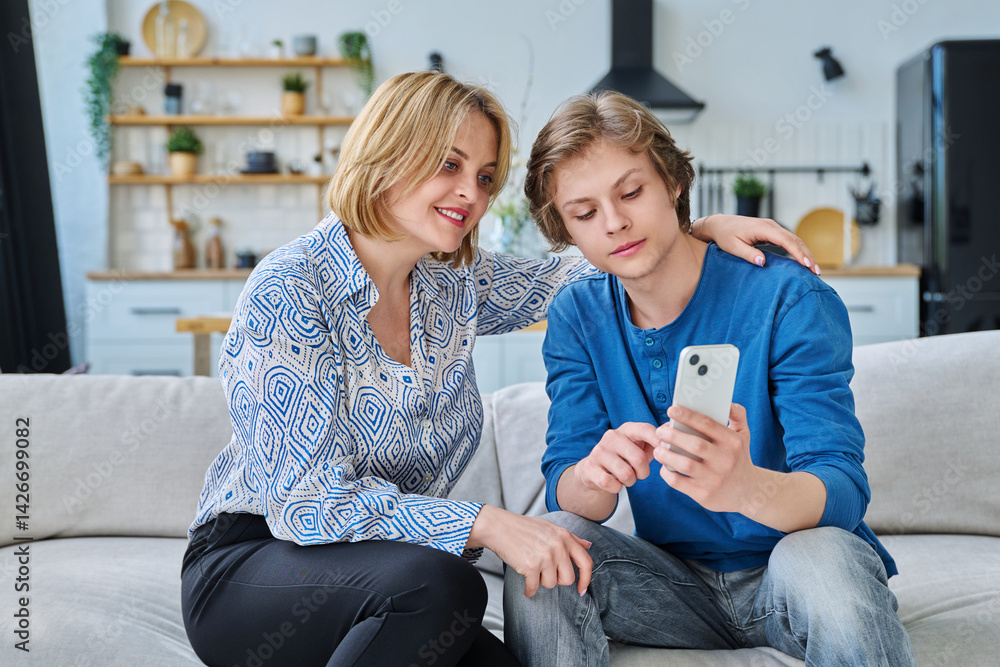 Middle aged mother and teenage son sitting together at home looking at smartphone