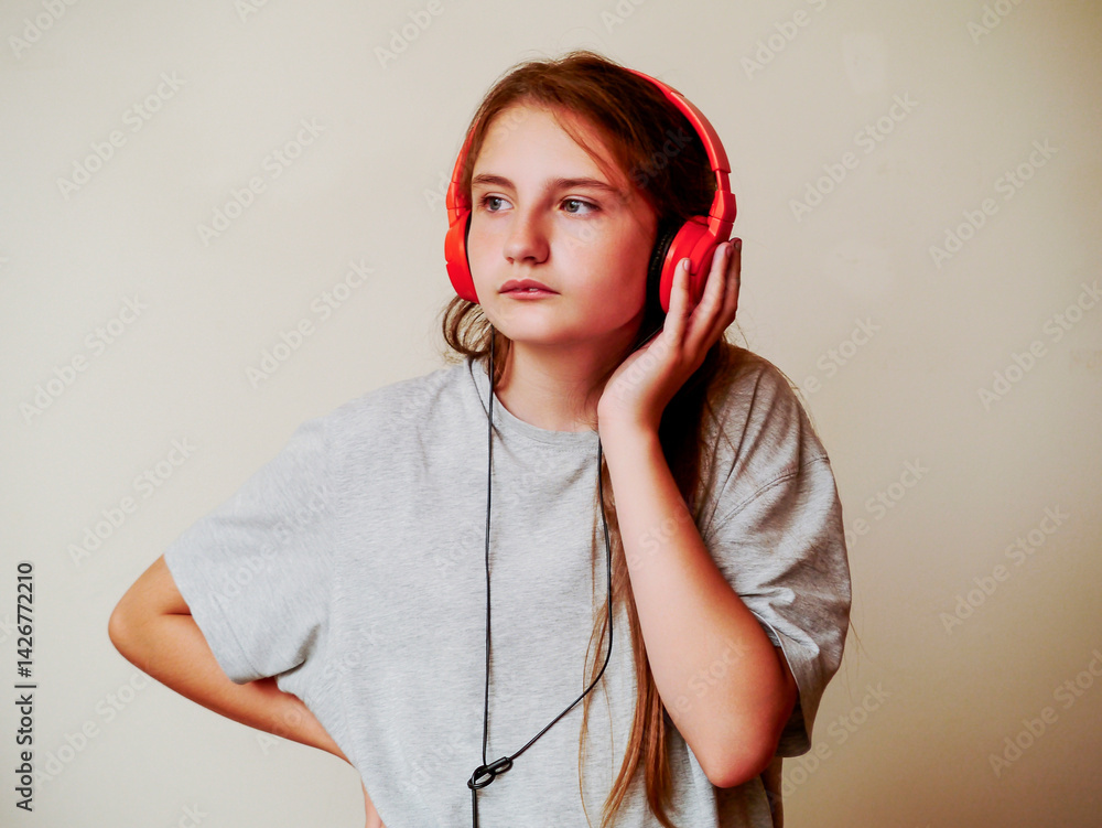Young girl in grey shirt listening music on red headphone. Selective focus. Future DJ. Young person enjoys audio. Light cream wall background.