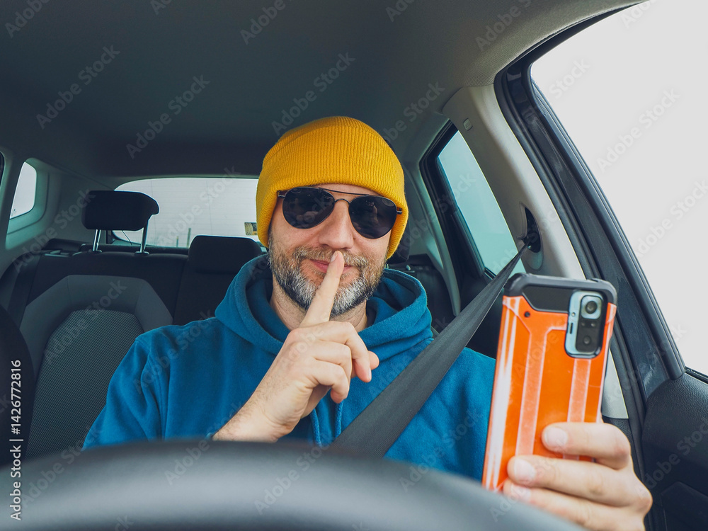 Man driver in blue jacket and yellow hat wearing dark aviator glasses holding smart phone. Travel and transport. Road safety concept. Male model in his 50s with grey beard dressed for outdoor trip.
